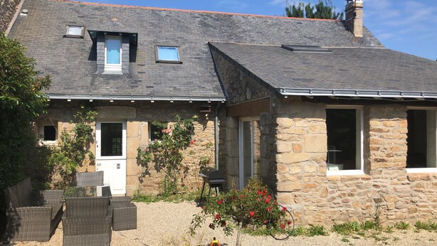 Une maison en pierre avec un toit en pente, deux fenêtres et un petit porche aménagé avec des meubles en osier.