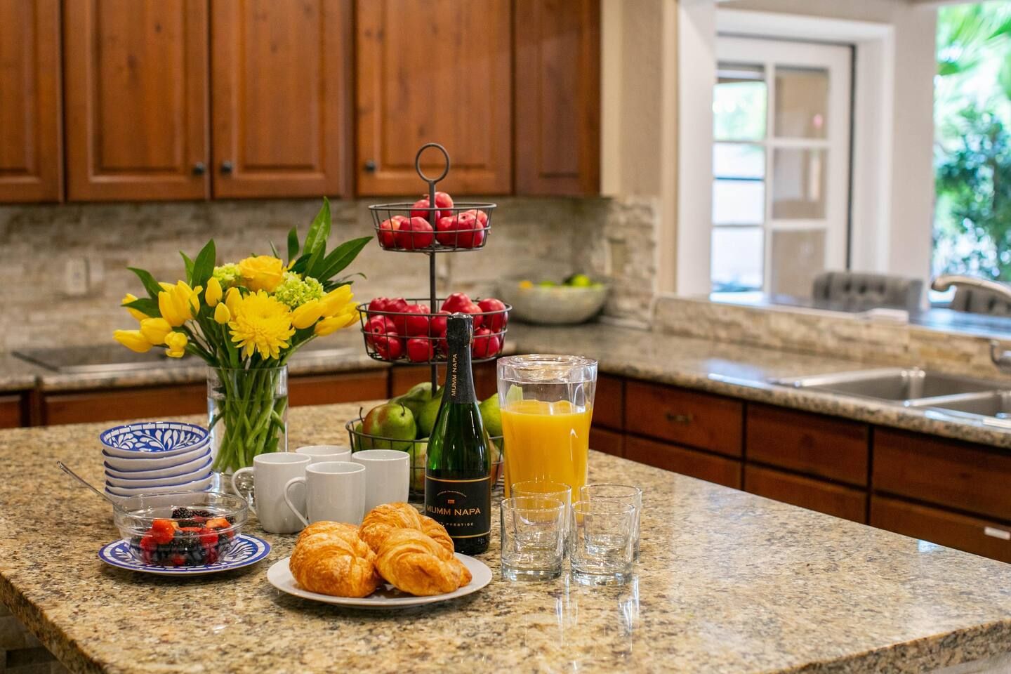A breakfast setting with croissants, a bottle of Mumm Napa champagne, fresh flowers, and a glass of orange juice on a granite countertop.