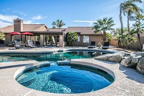 A swimming pool with a hot tub, lounge chairs, and palm trees.