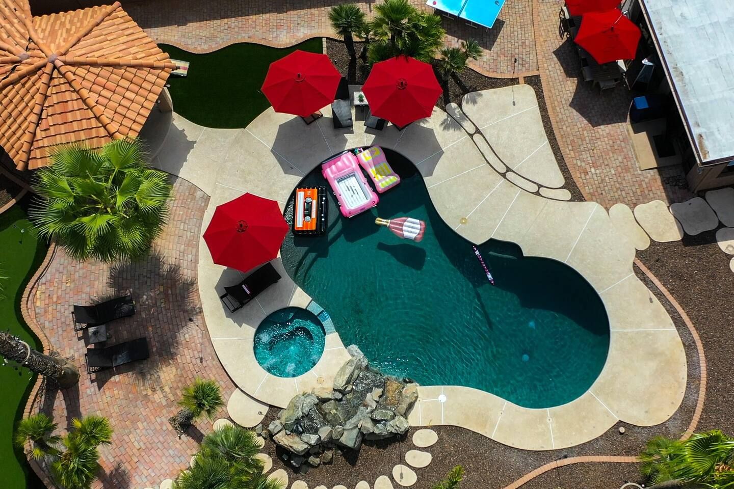 Vista aérea de un área de piscina con sillas de descanso, sombrillas y un jacuzzi pequeño.