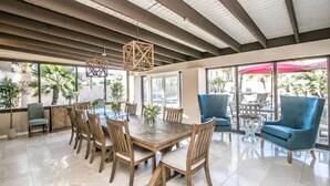 Un vaste espace dédié à la salle à manger, avec une grande table et des chaises en bois, un plafond avec des poutres apparentes et de grandes fenêtres offrant une vue sur l’extérieur.