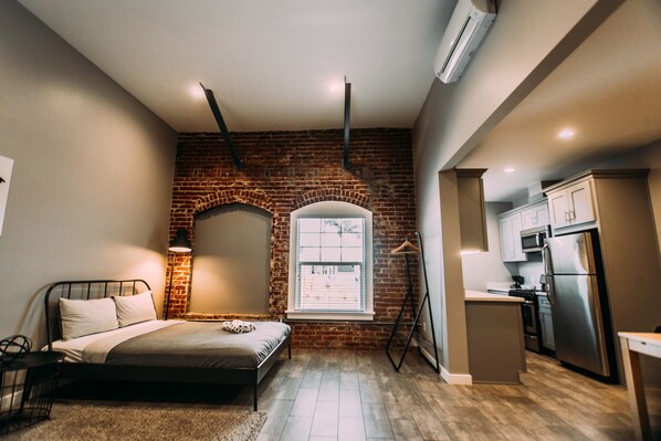 A bedroom with a brick wall, a bed, a nightstand, a lamp, and a window.