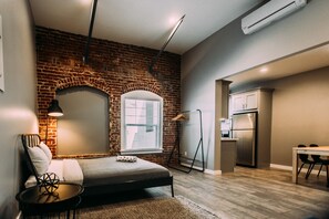 A modern bedroom with a brick wall, a bed, a desk, and a kitchenette.