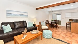 Ein Wohnzimmer mit einer schwarzen Ledersofa, einem Holztisch und einem Korbstuhl. Es gibt einen Essbereich mit Tisch und Stühlen sowie eine Küchenzeile mit Arbeitsfläche und Barhocker.