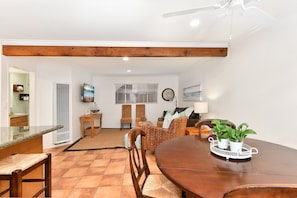 A dining area with a wooden table, chairs, and a tray with potted plants. There is a kitchen area with a refrigerator, stove, and sink in the background.