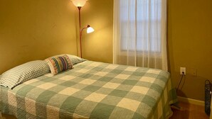 A bedroom with a bed, a lamp, a window with curtains, and a wall socket.