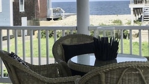 A covered outdoor seating area with wicker chairs and a table, overlooking a beach and a house. - Oceanfront Beach House/ Fall asleep listening to the waves (Scituate)