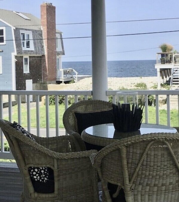Ein überdachter Außenbereich mit Korbstühlen und einem Tisch mit Blick auf Strand und Haus.