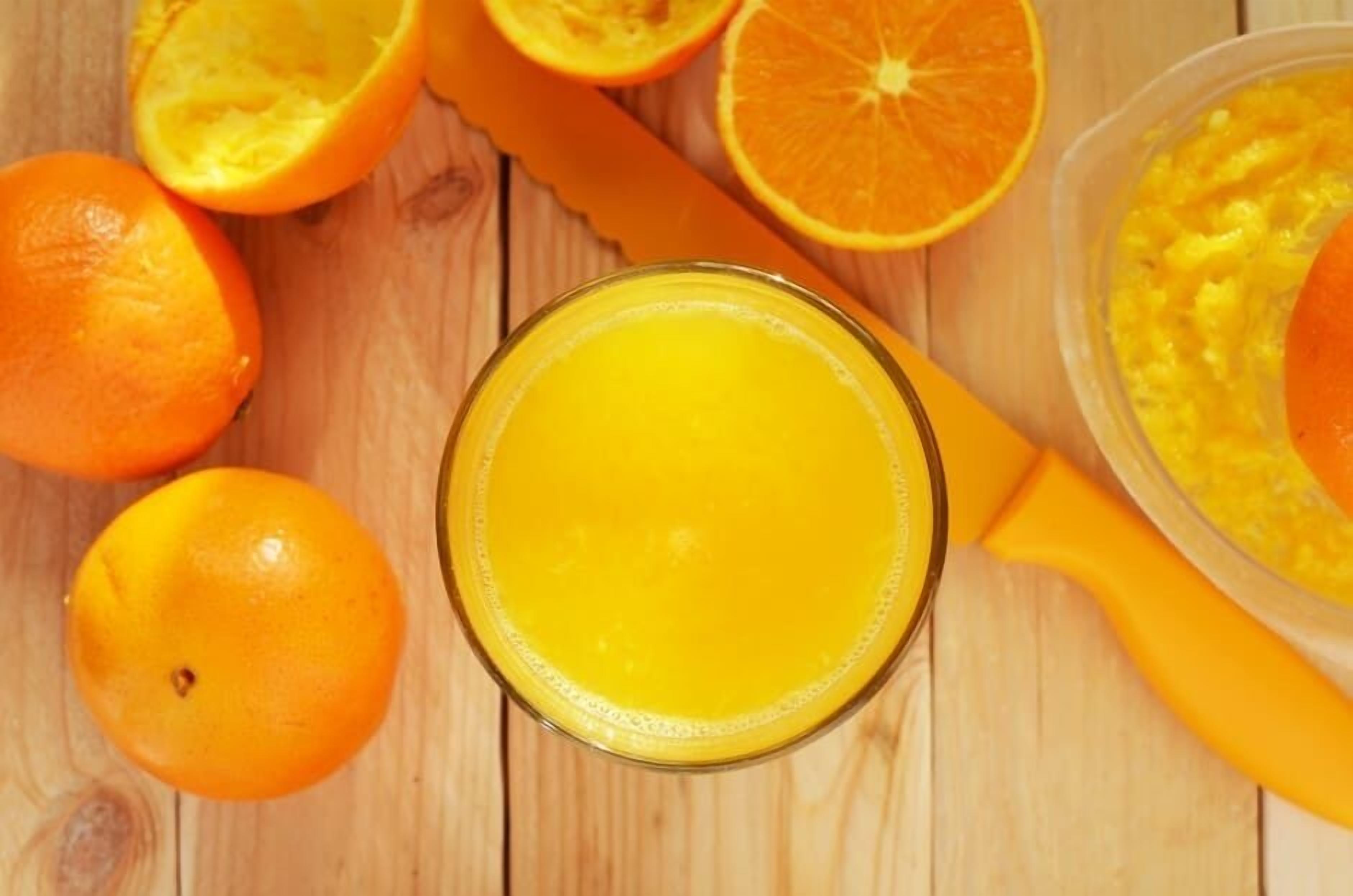 Un vaso de jugo de naranja sobre una mesa de madera, con naranjas enteras y cortadas alrededor.