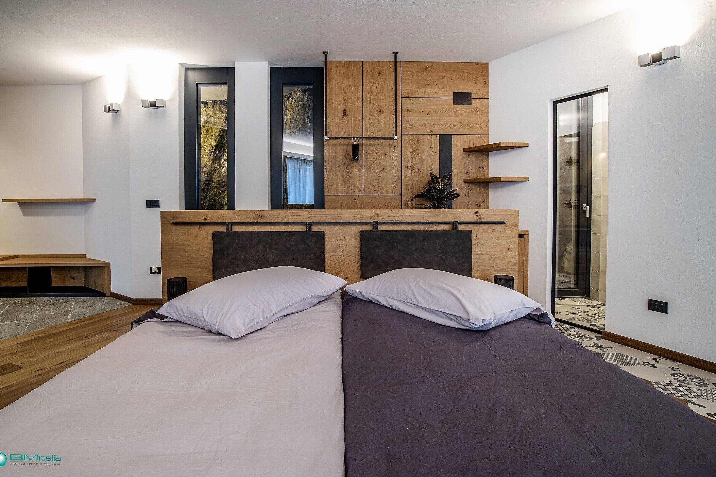 A modern bedroom with a bed, wooden headboard, built-in shelves, and a glass door leading to a bathroom.