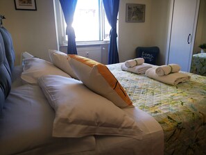A neatly made bed with white and orange pillows, a floral quilt, and rolled towels on a bed with a window view.