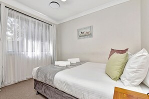 Un dormitorio con una cama, una ventana con cortinas translúcidas, un cuadro en la pared y suelo alfombrado.