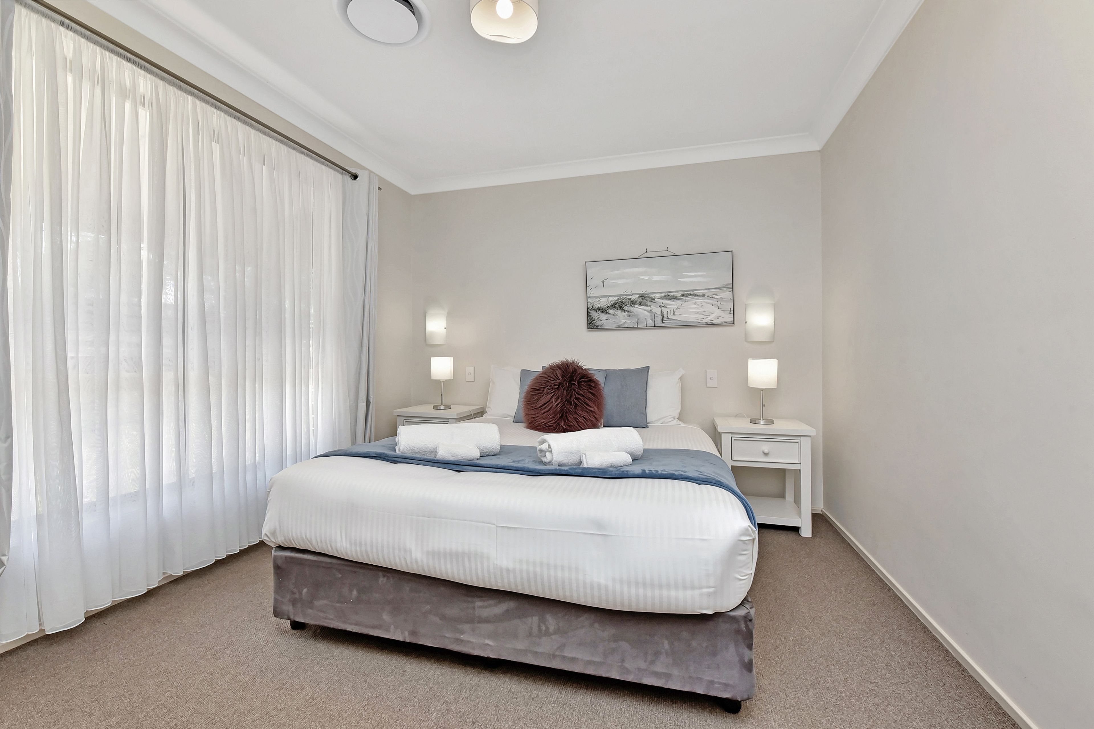 Une chambre moderne avec un grand lit, deux tables de chevet, une œuvre d’art accrochée au mur et une fenêtre ornée de rideaux légers.