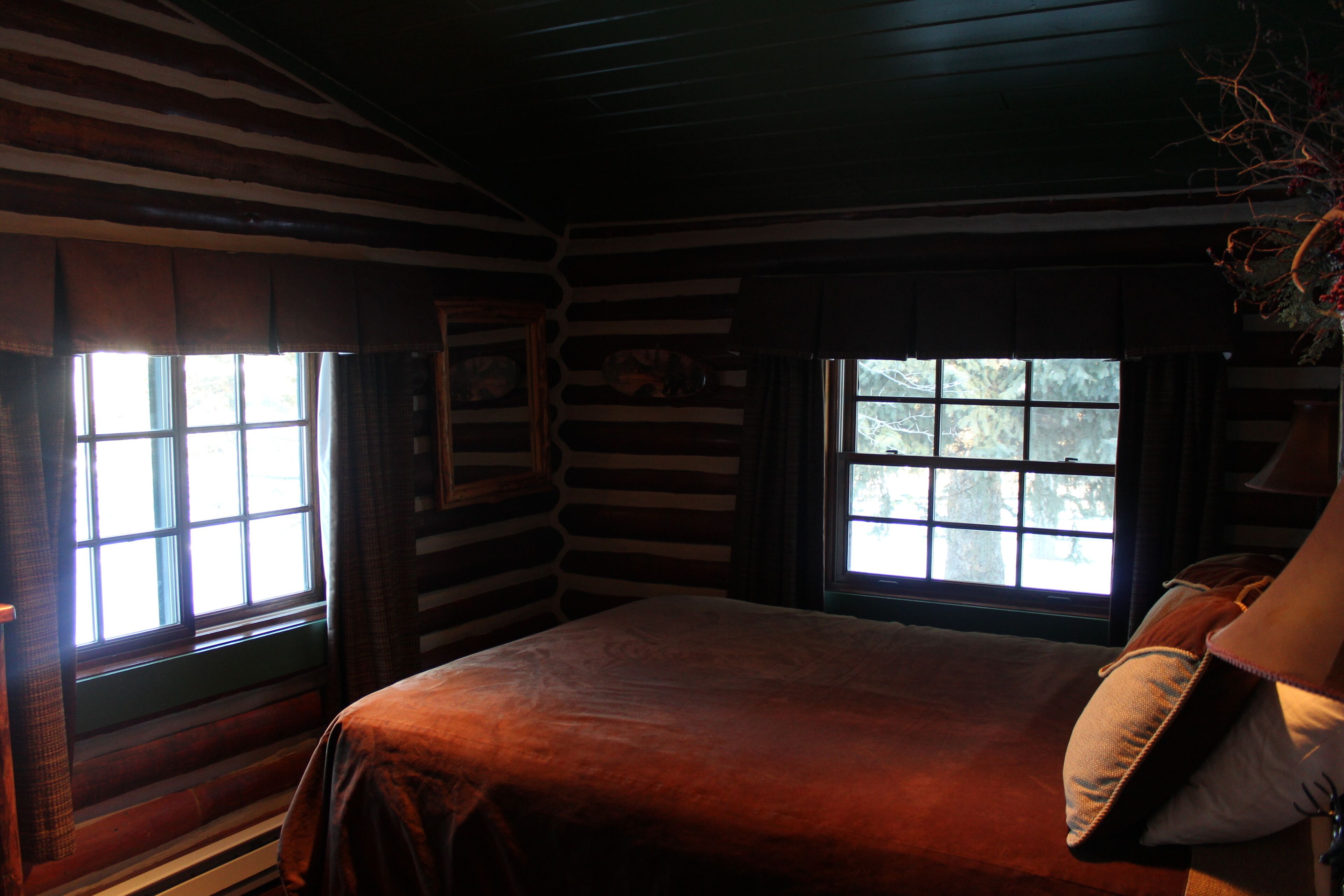 Habitación tipo cabaña de madera con una cama grande, paredes de madera y dos ventanas con cortinas.