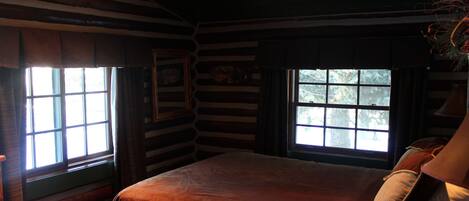 Habitación tipo cabaña de madera con una cama grande, paredes de madera y dos ventanas con cortinas.