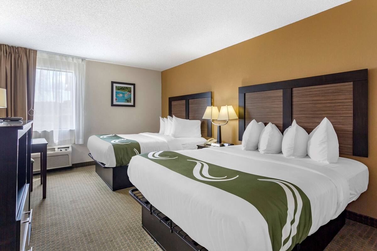 A hotel room with two beds, a desk, a chair, a lamp, and a picture on the wall.
