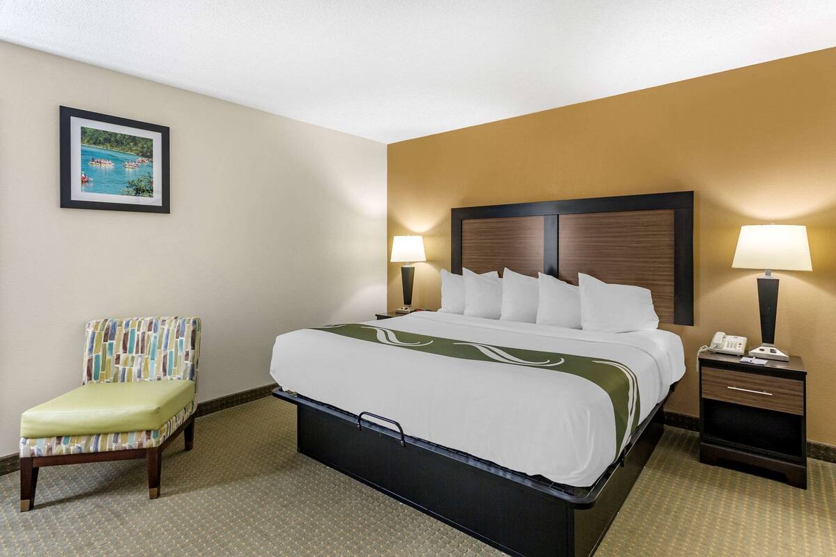 A hotel room with a bed, a chair, a nightstand, and a wall-mounted lamp.