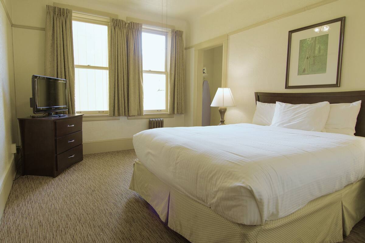 A hotel room with a large bed, a television on a dresser, a window with curtains, a lamp, and a framed picture on the wall.