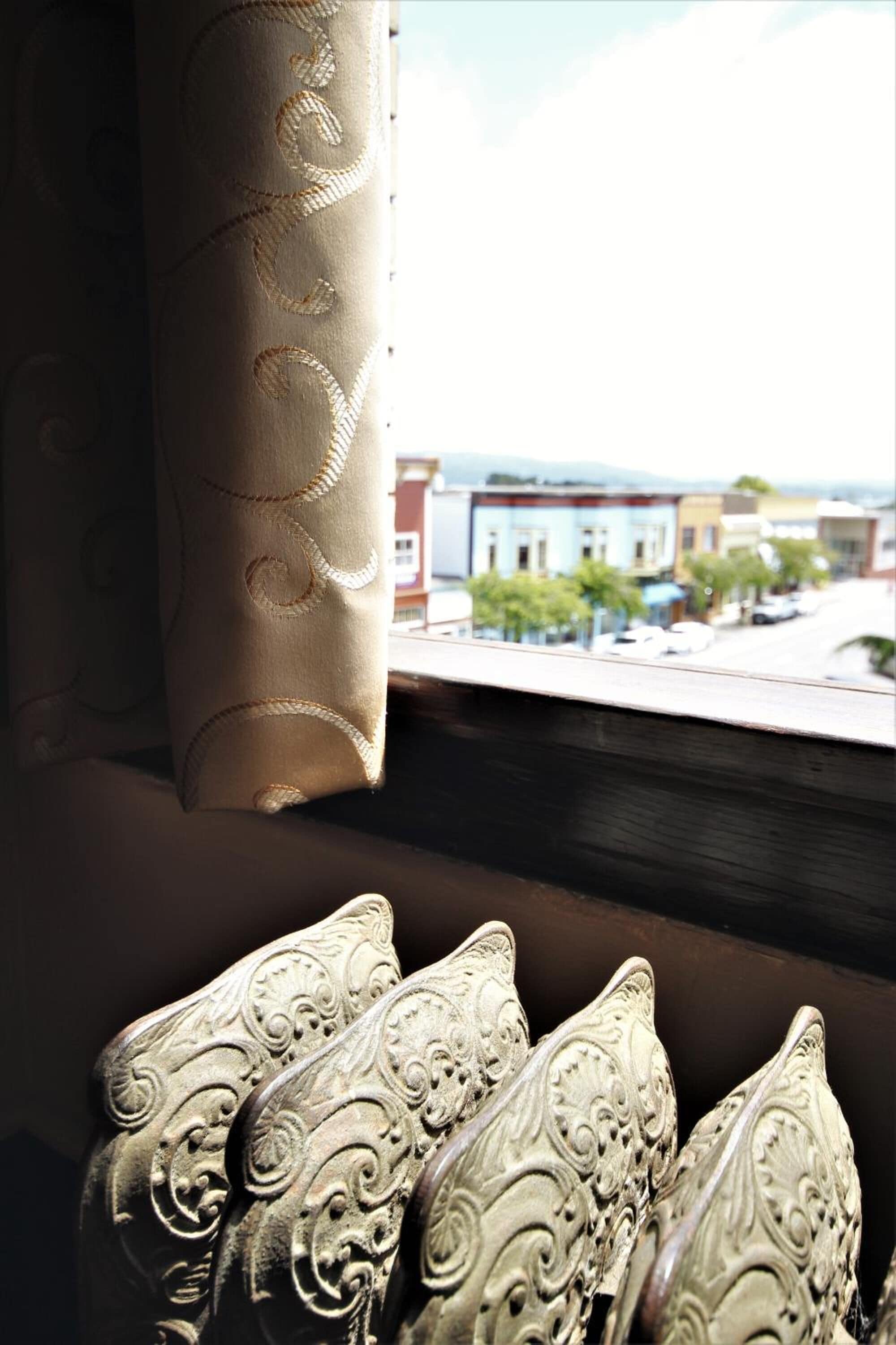 Una ventana con vistas a una calle y edificios, una cortina con un estampado, y un objeto decorativo con un diseño tallado.