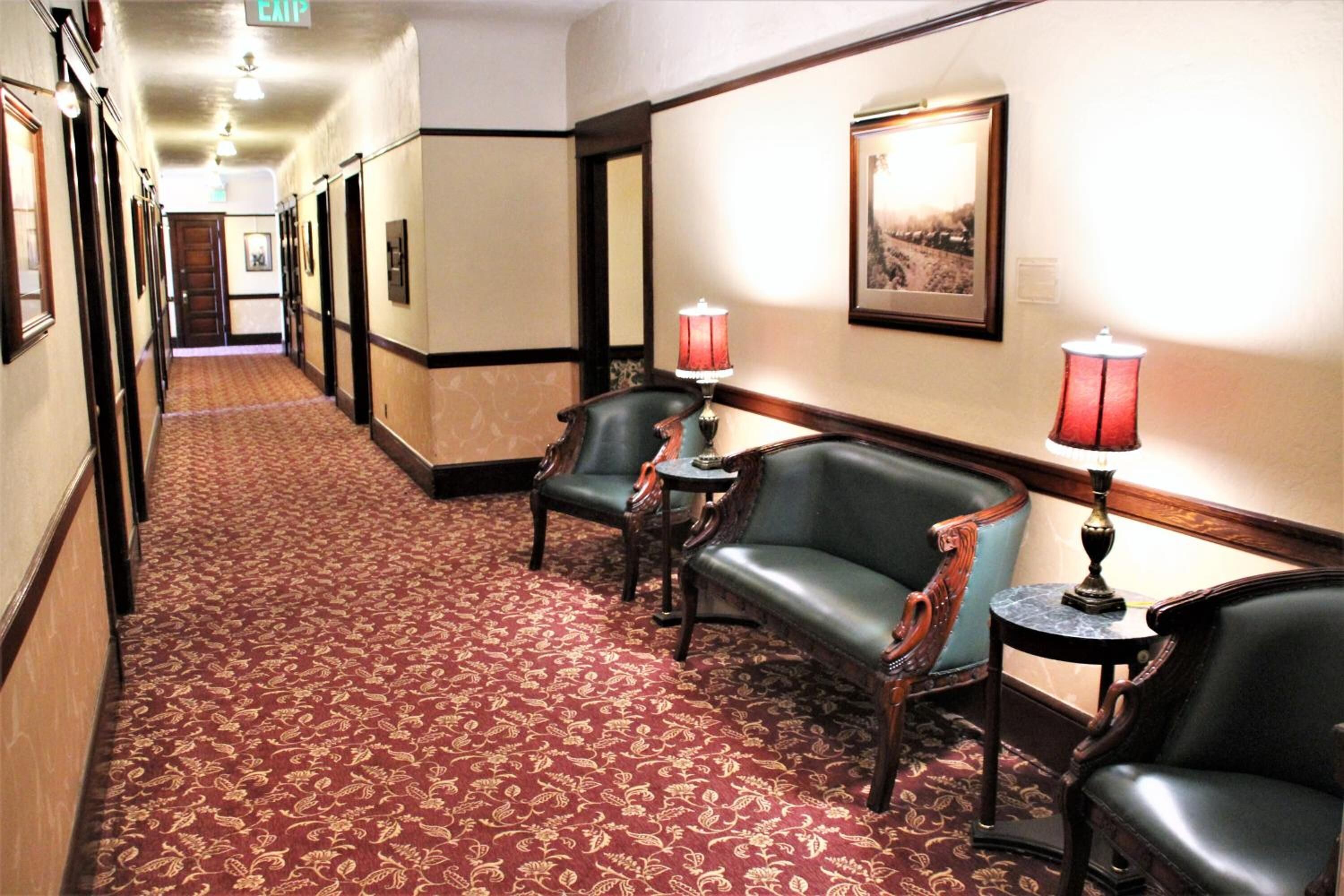 Un pasillo de hotel con alfombra estampada, sillas de madera y un cuadro enmarcado en la pared.