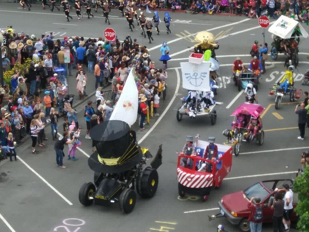 A parade with various floats and participants, including a large black vehicle and a red vehicle with a white and red design.