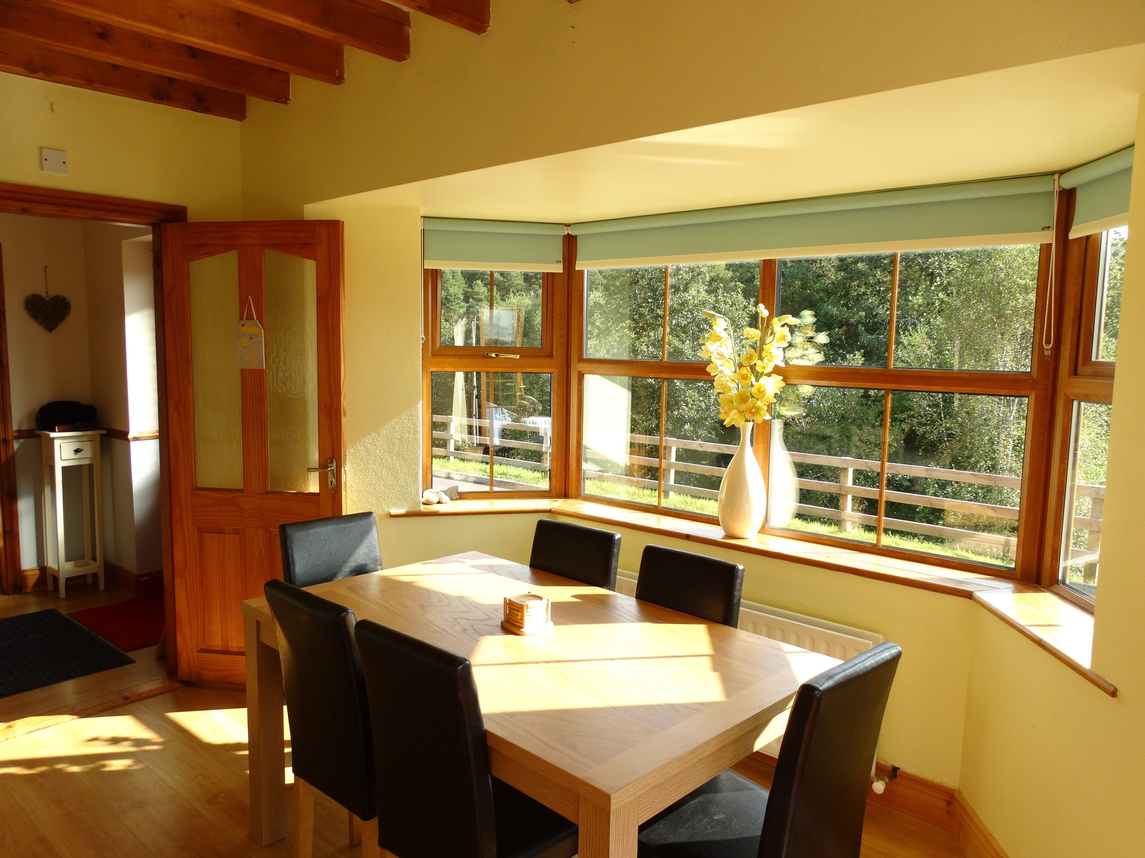 A dining area with a wooden table and chairs, a vase with flowers, and a view of greenery outside through large windows.