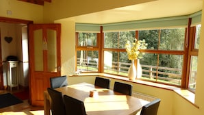 A dining area with a wooden table and chairs, a vase with flowers, and a view of greenery outside through large windows. - The River House, Nature Reserve Glengarriff (Glengarriff)