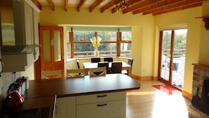 A kitchen with wooden beams, a dining area with a table and chairs, and a view through large windows. - The River House, Nature Reserve Glengarriff (Glengarriff)