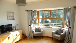 A cozy living room with a TV, two armchairs, a wooden cabinet, and a large window with curtains. - The River House, Nature Reserve Glengarriff (Glengarriff)