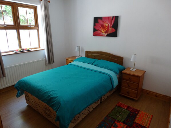 Une chambre à coucher avec un lit, deux tables de chevet, une commode, une fenêtre avec des rideaux, un radiateur et un tableau au mur.
