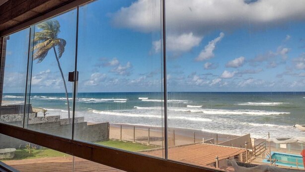 Vista desde un balcón sobre una playa con olas, una palmera y cielo despejado.