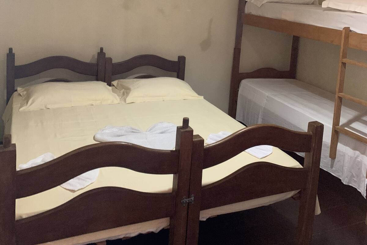 Uma cama de solteiro com estrutura de madeira e roupa de cama branca, uma cama beliche à direita e um ventilador de teto acima.