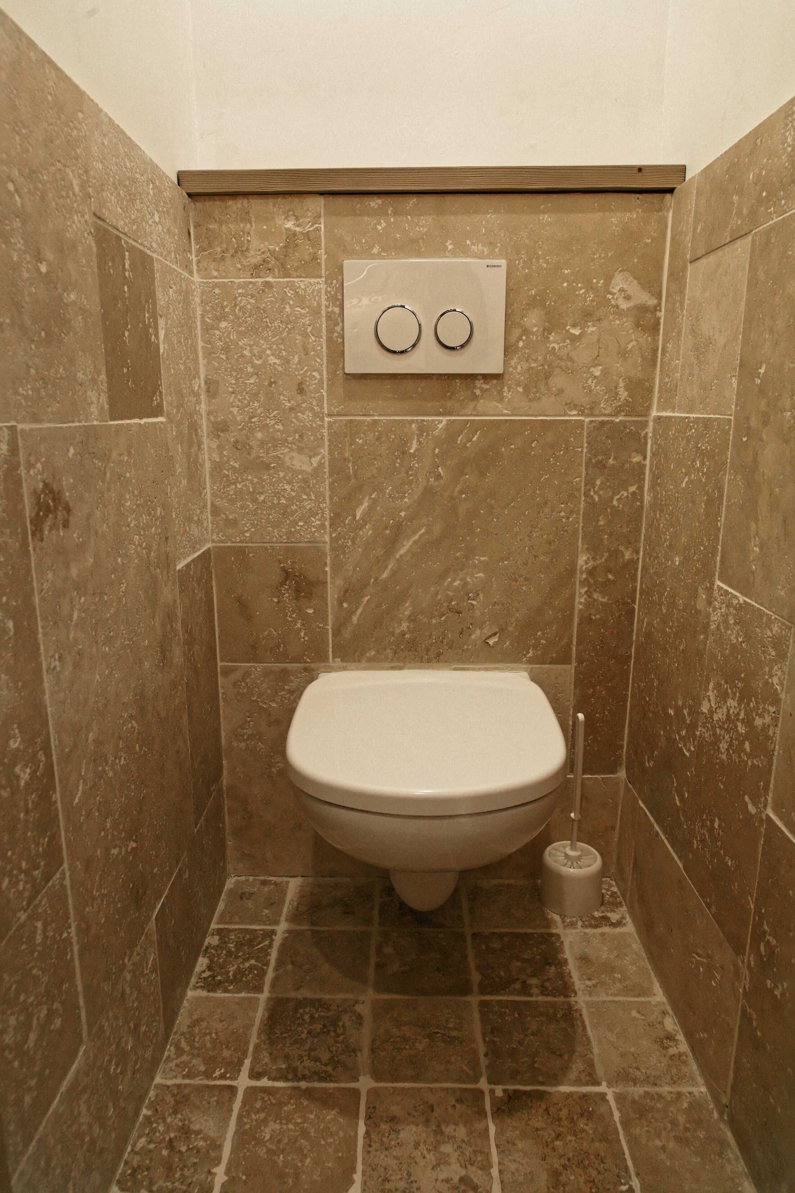 A small, tiled bathroom with a toilet, a flush handle, and a toilet brush.