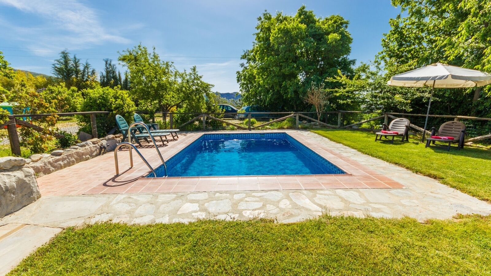Une piscine rectangulaire avec une terrasse carrelée, entourée d’une bordure en pierre et d’une zone herbeuse.