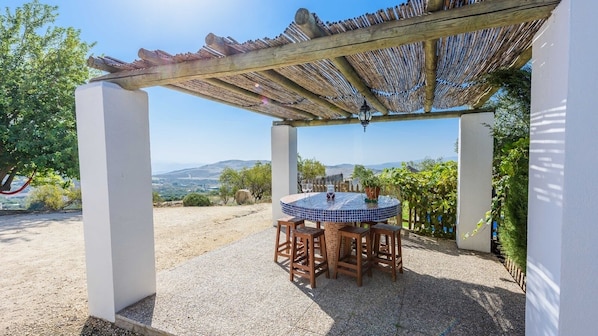 A covered outdoor dining area with a table and chairs, a pergola overhead, and a view of the landscape.