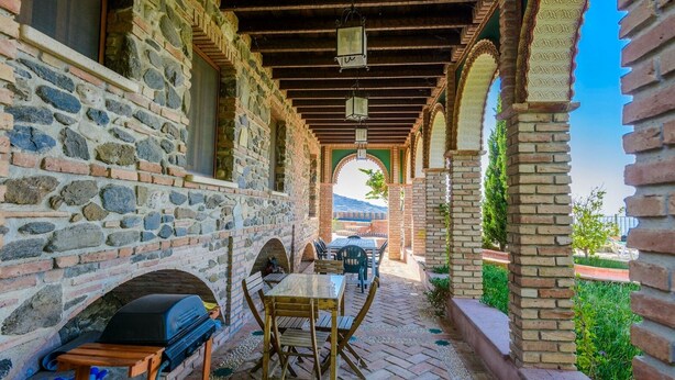 Eine Terrasse im Freien mit Steinwänden, einem Holztisch und -stühlen, einem Grill und Blick auf die Landschaft.