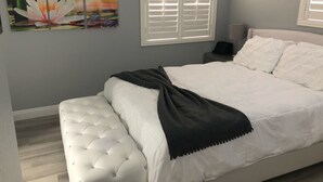 A bedroom with a white bed, a dark throw blanket, and a wall-mounted painting of a flower.