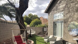 A backyard with a hammock, two wicker chairs, a small table, and a wall-mounted TV.