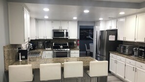 A modern kitchen with white cabinets, stainless steel appliances, and a granite countertop. There are three bar stools and a dining area visible.