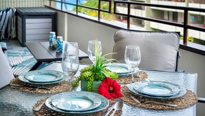 Mesa de comedor dispuesta con platos azules, vasos transparentes y un centro de mesa con plantas verdes y una flor roja, con vistas a un balcón que da a edificios y vegetación.