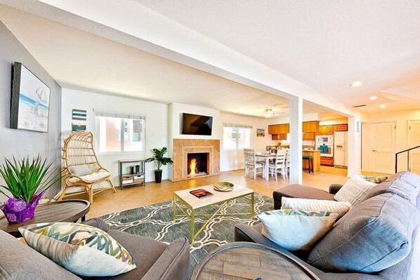 A spacious living room with a fireplace, comfortable seating, and a kitchen area visible through an open door.
