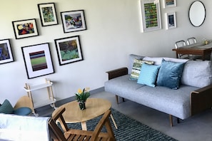 A living room with a grey sofa, wooden coffee table, and a small round table with flowers. There are framed pictures on the wall and a dining table with chairs.