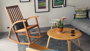 Ein Wohnzimmer mit einem Holzschaukelstuhl, einem kleinen runden Tisch mit einem Topfpflanzen und einem grauen Sofa. An der Wand befinden sich mehrere gerahmte Kunstwerke.