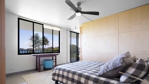 Ein Schlafzimmer mit Bett, Deckenventilator, Schreibtisch und großem Fenster mit Blick auf Palmen.
