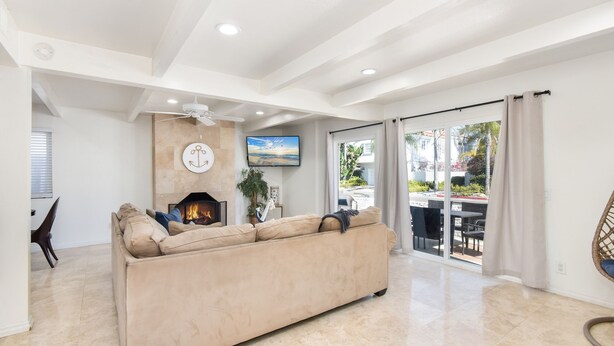 A spacious living room with a beige sofa, a fireplace, a ceiling fan, and a large window with curtains.
