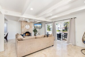 Een ruime woonkamer met een beige bank, een open haard, een plafondventilator en een groot raam met gordijnen.