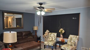 Ein Wohnzimmer mit einem braunen Ledersofa, zwei gemusterten Sessel, einem Deckenventilator, einem großen Spiegel und einer Tafel an der Wand.
