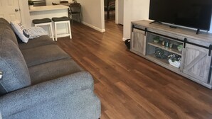 Ein Wohnzimmer mit grauem Sofa, einem Fernseher auf einem Holzschrank und einer Küche im Hintergrund.