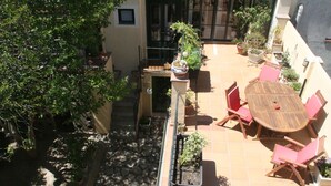 Un patio con una mesa redonda de madera y sillas rojas, rodeado de plantas en macetas y un árbol.