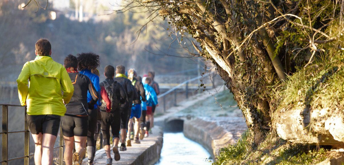 A group of people jogging along a riverside path with a stone wall on one side and trees on the other.